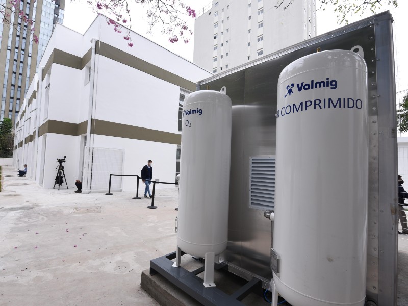 Foto da miniusina de oxigênio instalada. Do lado direito aparece uma estrtura metálica quadrada com dois cilindros brancos grandes acoplados. Ao fundo está o prédio branco da futura UPA Vila Mariana e mais para trás aparecem outros dois prédios